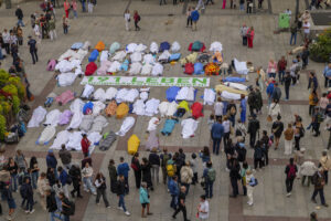 Das Bild zeigt die Aktion am Münchner Marienplatz zum Welttag der Suizidprävention am 10.09.2024. Man sieht Menschen, die mit Tüchern abgedeckt auf dem Boden liegen und ein Plakat mit der Aufschrift "191 Leben, Suizidprävention stärken!"