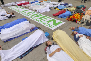 Das Bild zeigt die Aktion am Münchner Marienplatz zum Welttag der Suizidprävention am 10.09.2024. Man sieht Menschen, die mit Tüchern abgedeckt auf dem Boden liegen und ein Plakat mit der Aufschrift "191 Leben, Suizidprävention stärken!"